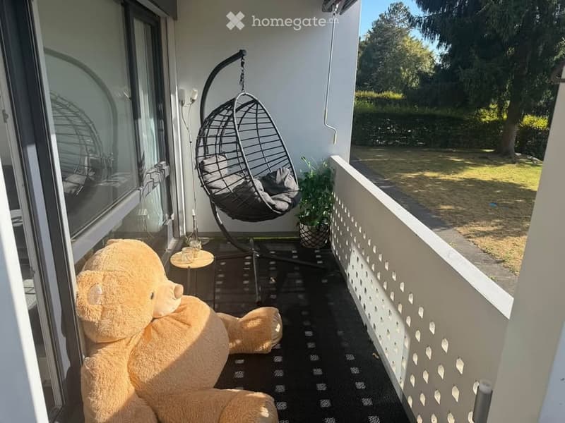 Balcony with a large teddy bear, a hanging wicker chair, a small table with a cup, and a potted plant. The balcony has a view of a green lawn and trees.