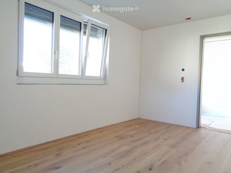 Empty room with light wooden flooring and white walls, featuring a large window with white frames and a doorway leading to another space.
