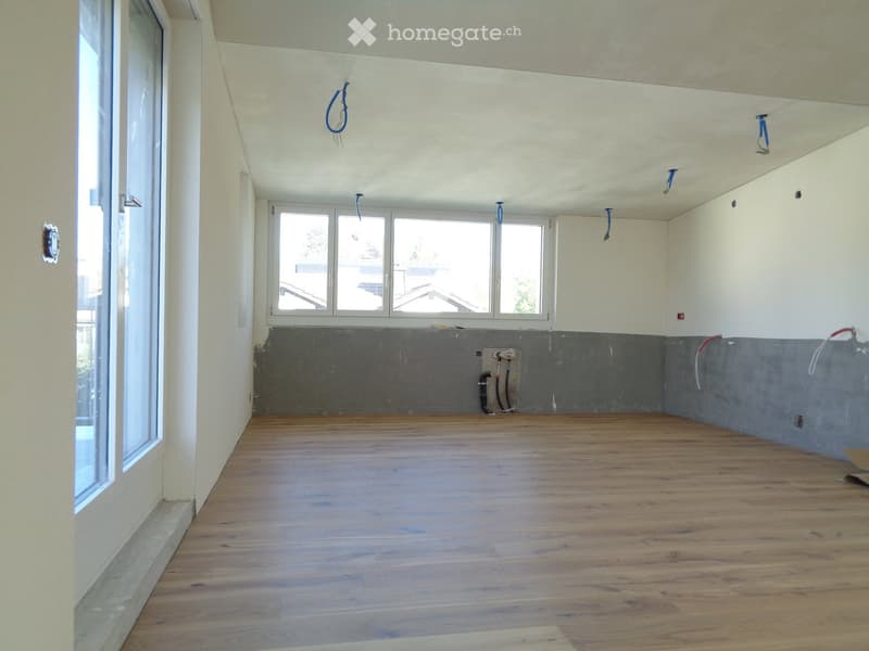 Empty room with wooden flooring, white walls, and large windows, showing exposed pipes and electrical outlets.