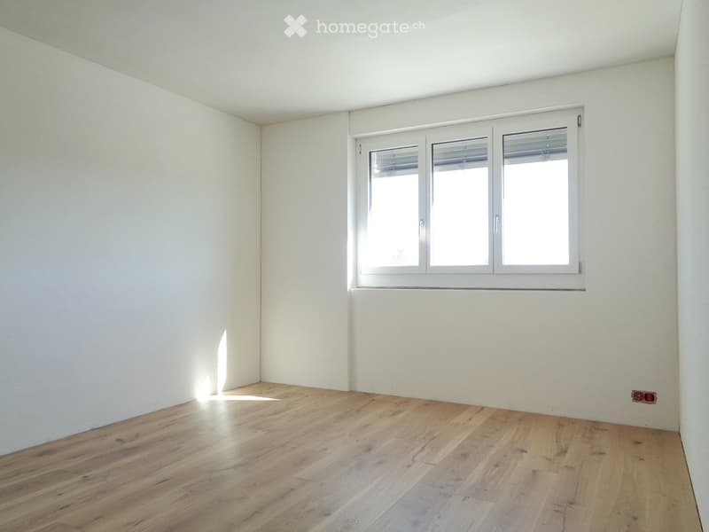 Empty room with light wooden flooring and white walls, featuring a large window with blinds allowing natural light to enter.