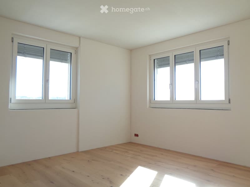 Empty room with wooden flooring and white walls, featuring multiple windows with blinds and a visible electrical outlet on the wall.