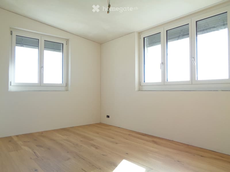 Empty room with light-colored wooden flooring and white walls, featuring multiple windows with white frames and blinds.