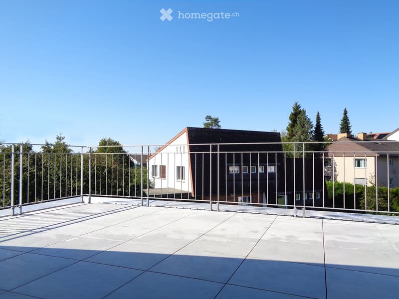 A modern rooftop terrace with a metal railing, tiled floor, and a view of surrounding buildings and trees.