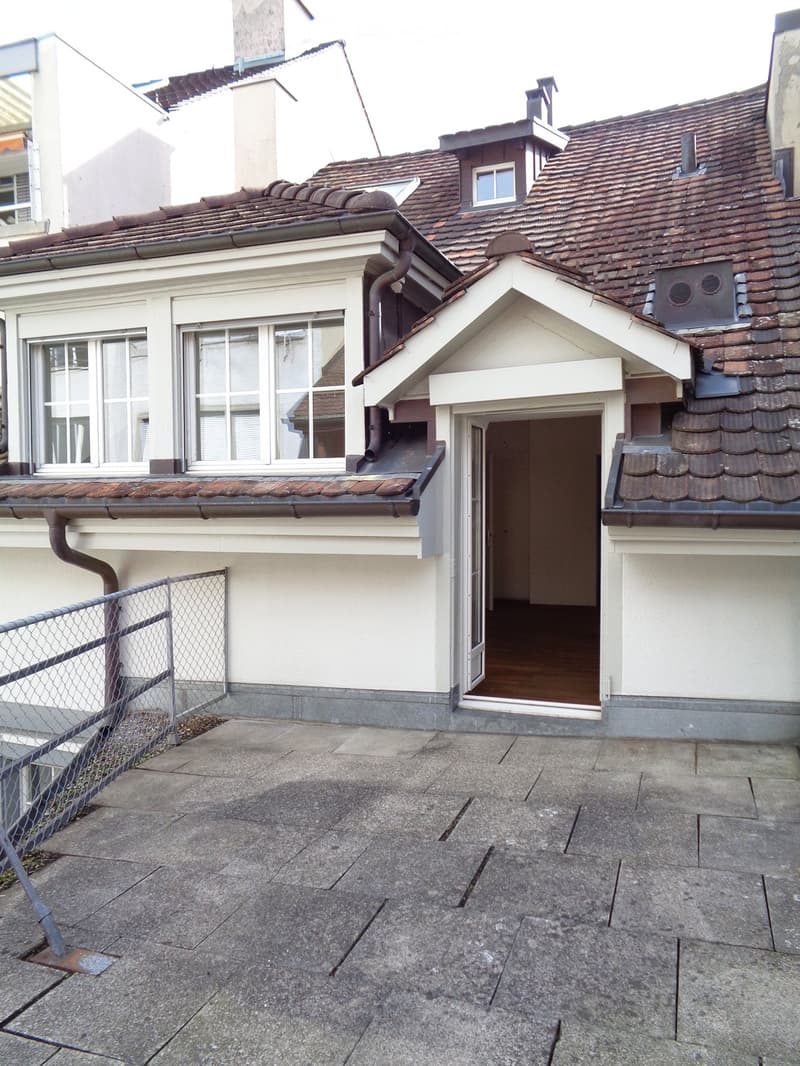 Roof terrace of a multi-story building with a concrete floor, metal fence, and access to a room with wooden flooring.