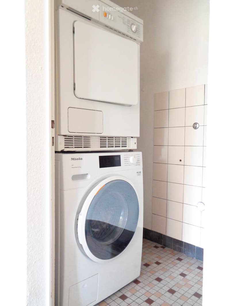 A compact bathroom with a white washing machine and dryer stacked vertically, beige tiled walls, and a small tiled floor.