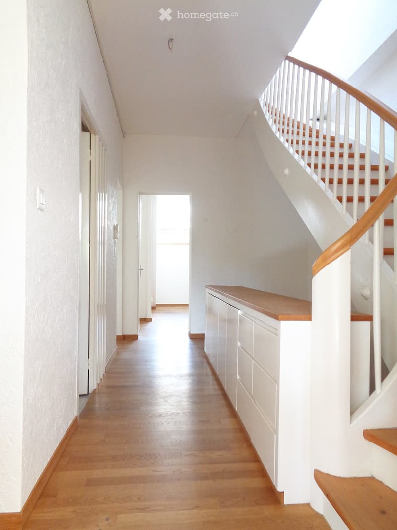 Bright hallway with wooden flooring, white walls, and a staircase leading upwards with wooden steps and a white railing. A small built-in cabinet is visible on the right side.