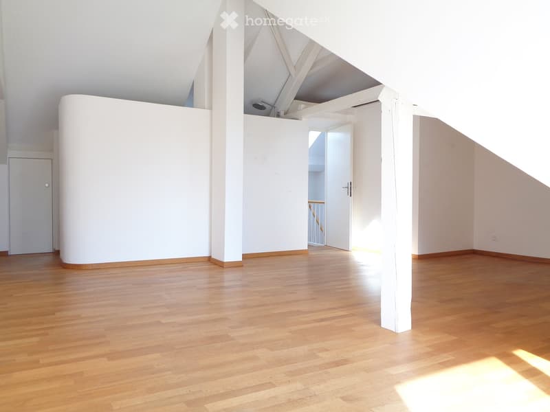 Empty room with wooden flooring, white walls, and a large open space. Features include a white column, a door, and a visible staircase in the background.