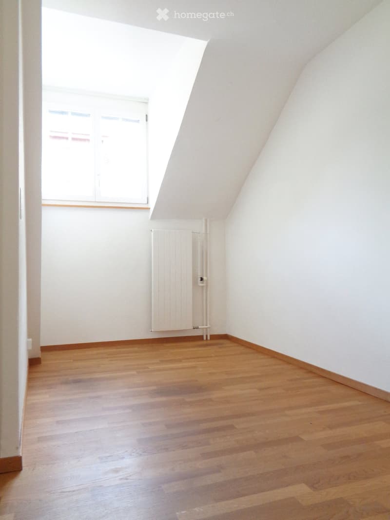 Empty attic room with wooden flooring, white walls, and a radiator. The room features a skylight and a window with blinds.