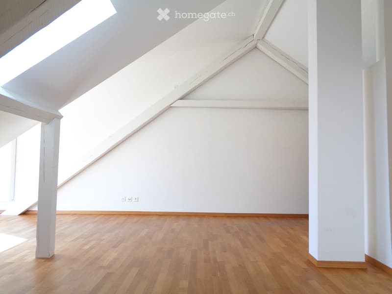 Empty attic room with wooden flooring, white walls, and exposed wooden beams. Natural light streams in from skylights.