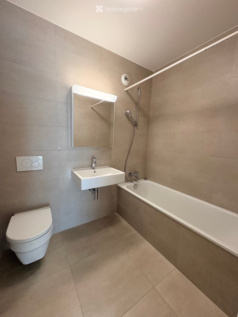 Modern bathroom with beige tiles, a toilet, a sink with a mirror, and a bathtub with a handheld shower.