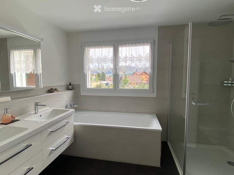 Modern bathroom with a double sink, bathtub, large mirror, and glass-enclosed shower. The room features light-colored tiles and a view of the neighborhood through the window.