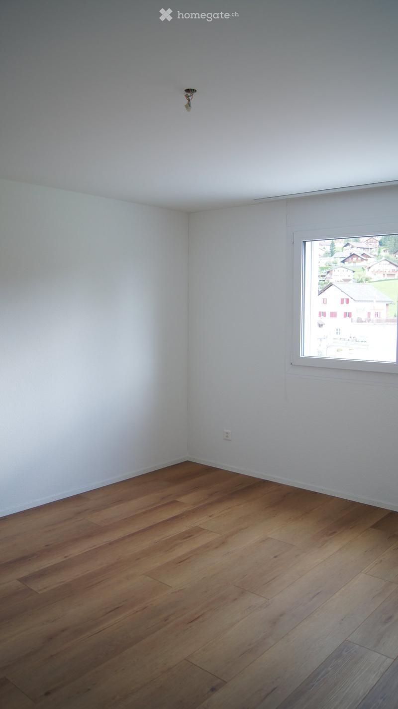 Empty room with light-colored walls and wooden flooring, featuring a single window with a view of the outside and a light fixture on the wall.