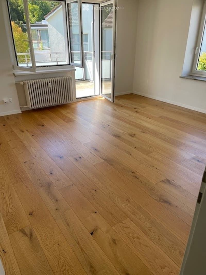 Empty room with light wooden flooring, large windows, and a radiator. No furniture or equipment present.