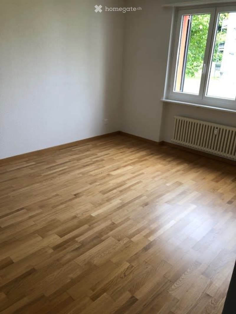 Empty room with wooden flooring, white walls, and a window with a radiator below it.