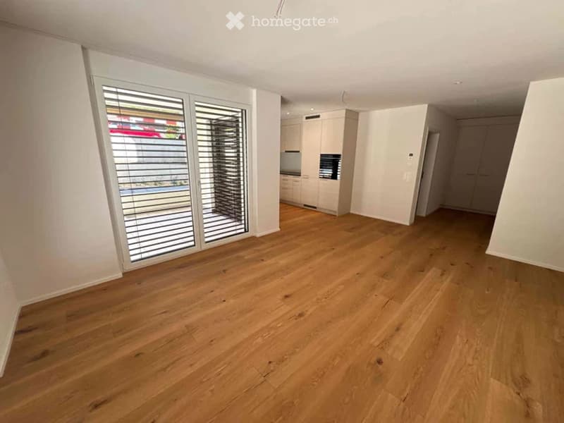 Empty room with light wooden flooring, large window with blinds, and a view of a car and outdoor area.