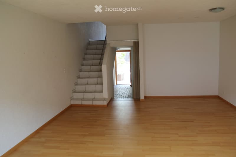 Empty room with wooden flooring, white walls, and a staircase leading upwards. A doorway leads to an outdoor area with a paved surface.