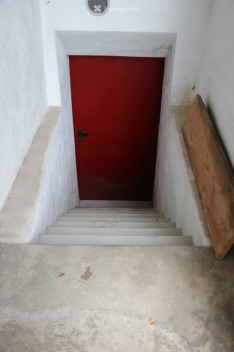 A narrow, white-walled corridor with a red door at the end and a concrete floor. A wooden plank leans against the wall to the right.