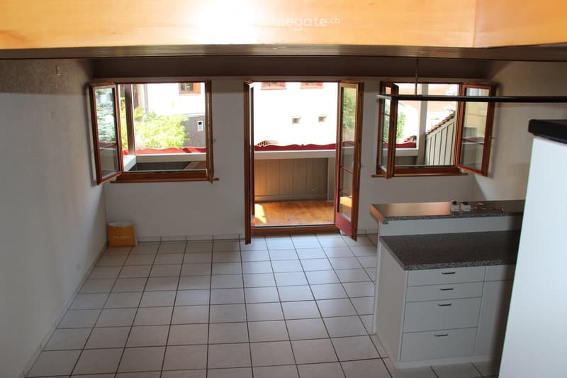 Empty room with tiled floor, white walls, and a small kitchen area with cabinets and a countertop. Large windows open to a balcony area with wooden flooring.