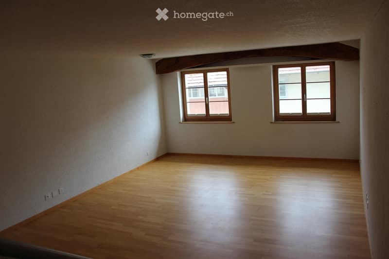 Empty room with wooden flooring and large windows, featuring exposed wooden beams on the ceiling.