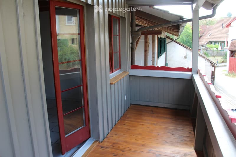 Wooden balcony with grey siding, red door, and small wooden shelf on the wall.