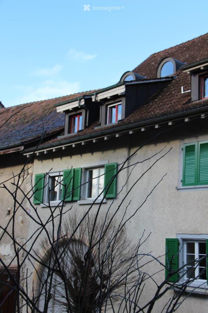 A multi-story building with a tiled roof, green shutters, and multiple windows. The facade is light-colored with a classic architectural style.