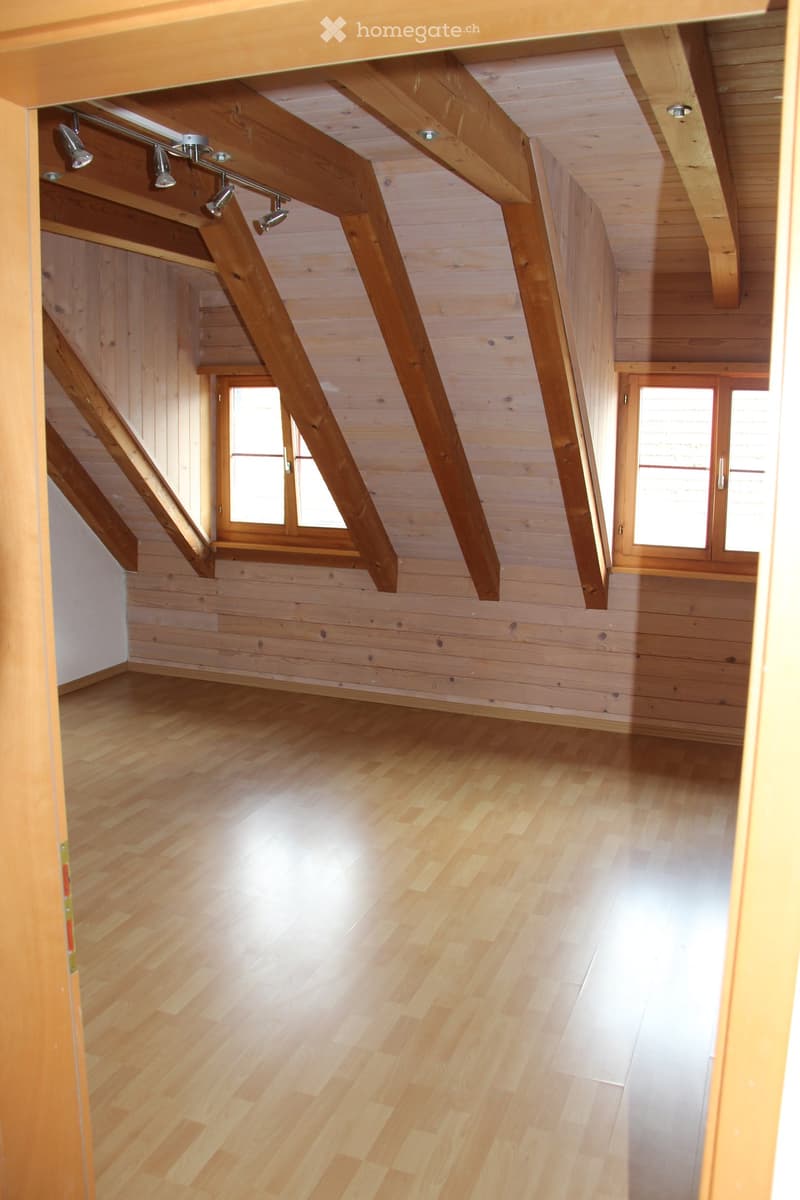 Empty room with wooden walls, wooden floor, and exposed wooden beams on the ceiling. Two windows with wooden frames are visible.