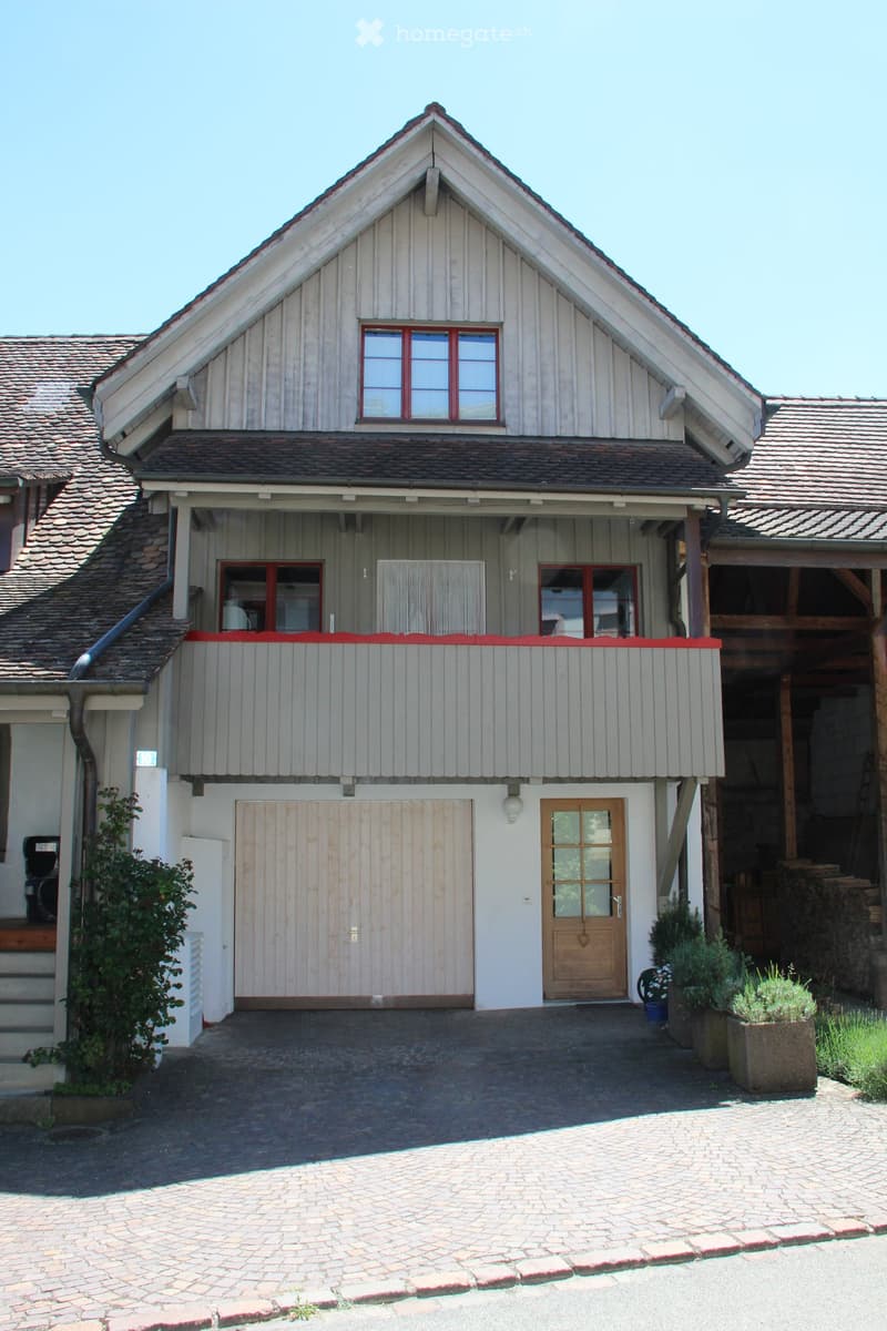 Two-story house with a gabled roof, wooden siding, and red-framed windows. Features a covered balcony area, a garage, and a paved driveway.