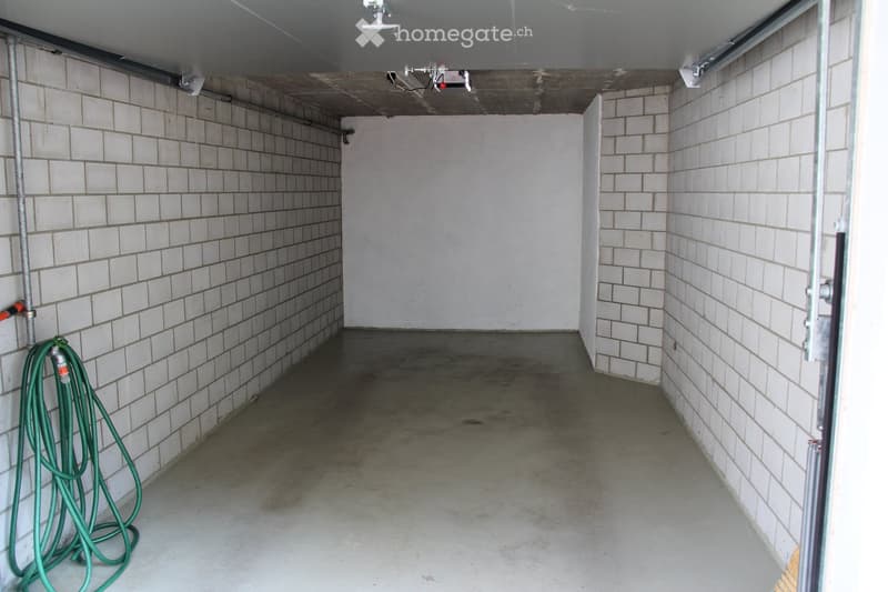 Empty room with white tiled walls and a concrete floor, featuring a coiled green hose on the left side and a partially open door on the right.