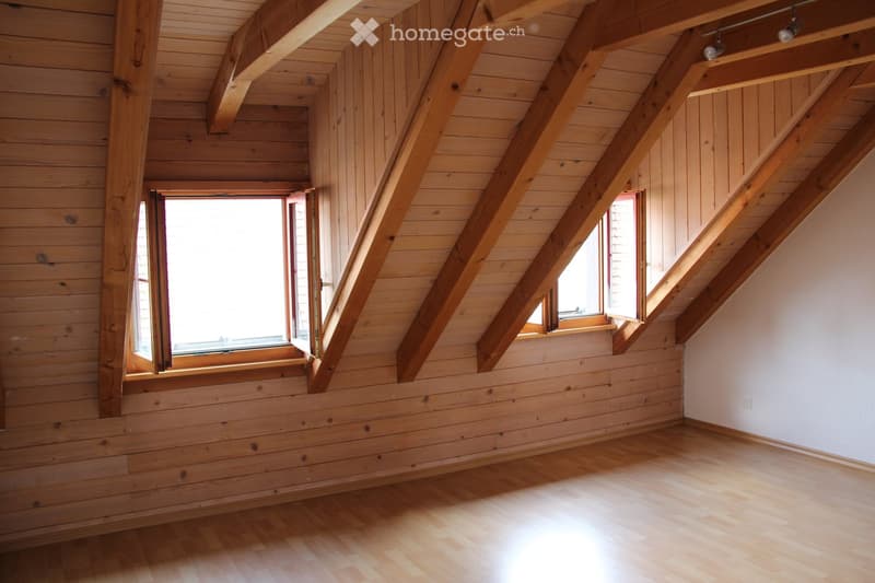 Empty attic room with wooden walls, wooden floor, exposed wooden beams, and two windows letting in natural light.