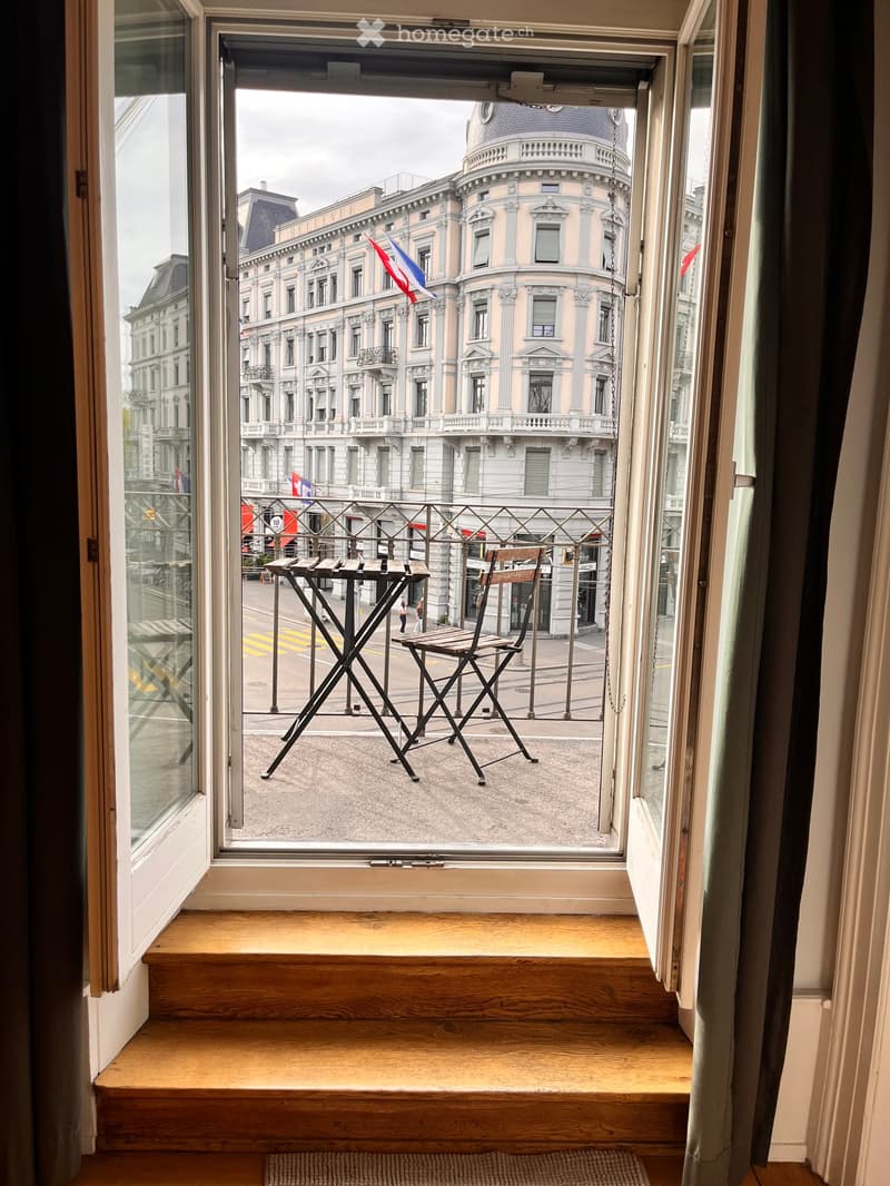 Balcony with a small table and chair, overlooking a street with historic buildings and flags.