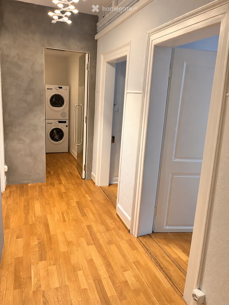 A narrow hallway with wooden flooring, white walls, and a modern light fixture. A door on the left leads to a room with a washing machine and dryer, while another door on the right is partially open.