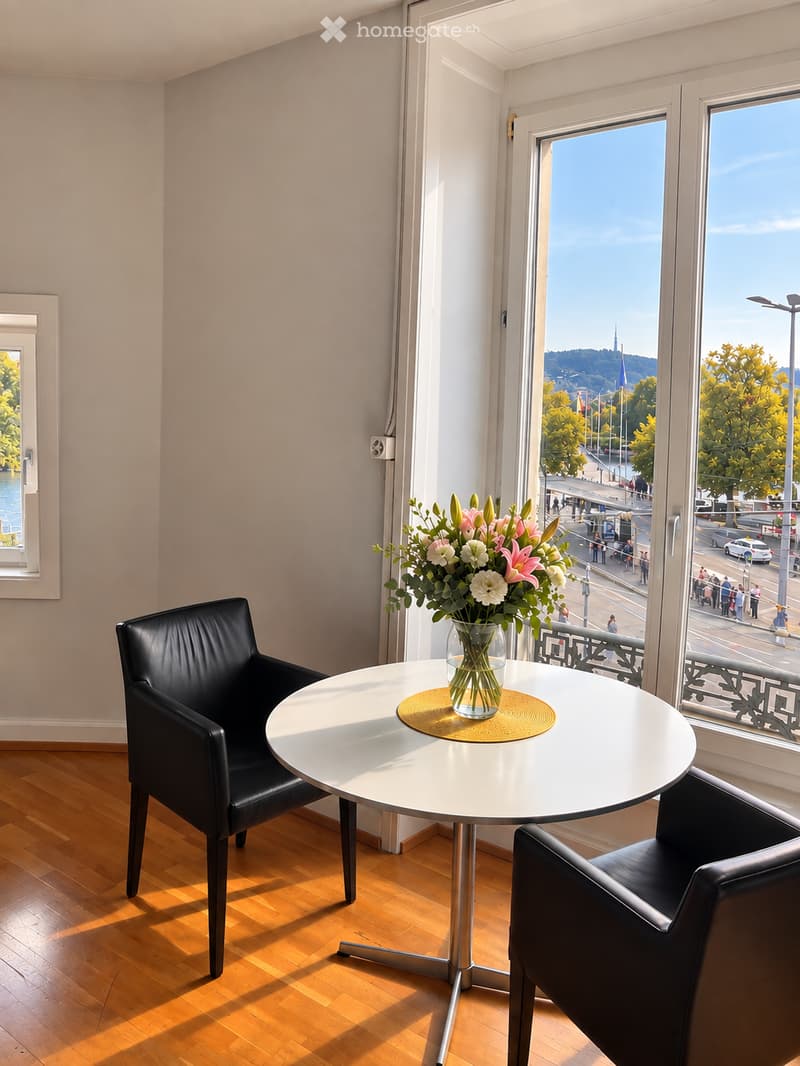 A small, modern room with wooden flooring, a round white table, two black leather chairs, and a vase of flowers on the table. Large windows offer a scenic view of the city and water.