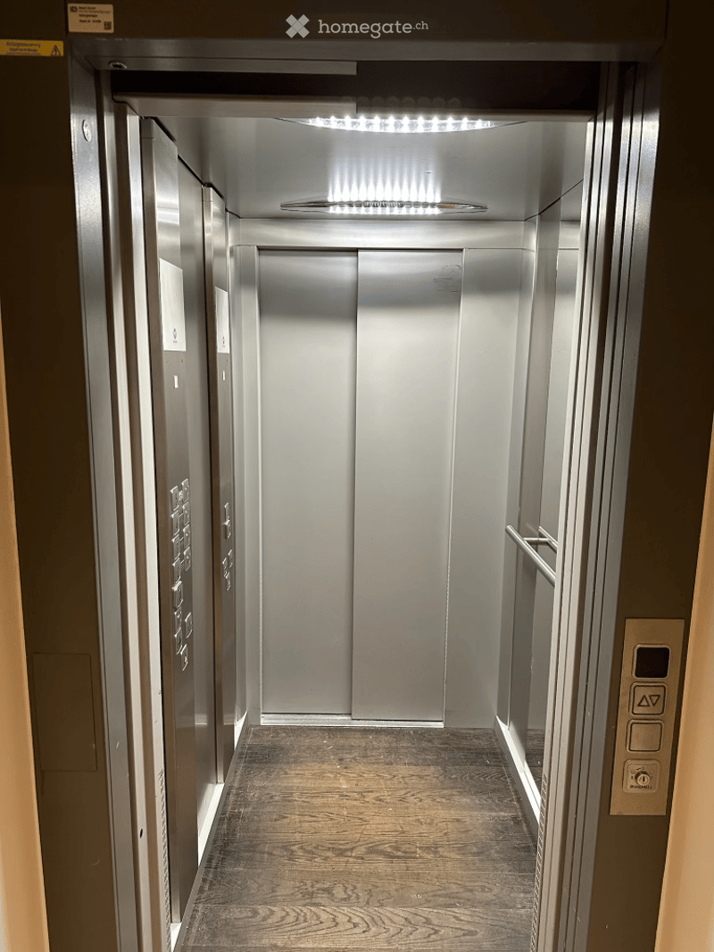 Modern hallway with wooden flooring, stainless steel elevator doors, and illuminated ceiling lights.