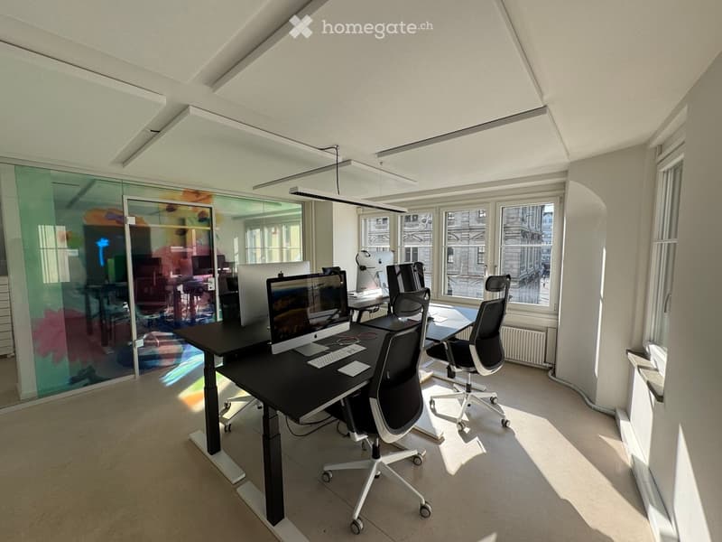 Modern office space with large windows, sleek black desk, ergonomic chairs, and multiple computer monitors.
