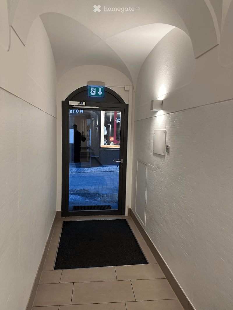 A modern hallway with arched ceiling, tiled floor, and a glass door leading outside. The door has a sign indicating an exit and a reflection of a person taking a photo.
