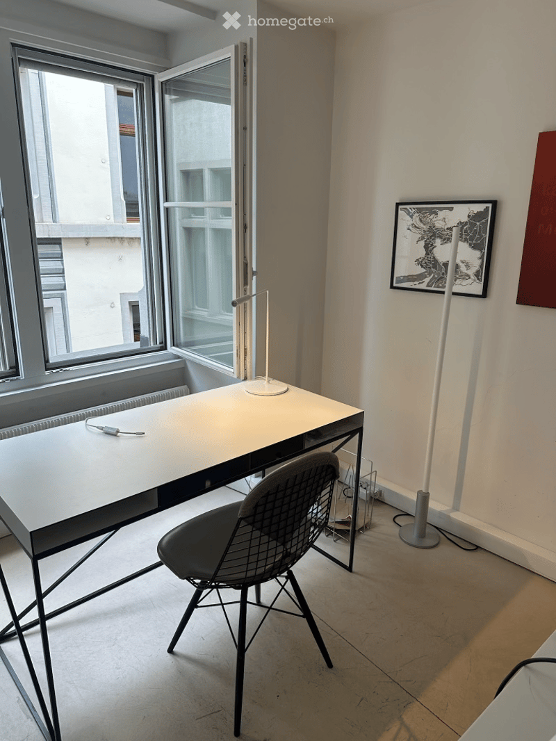 Modern, minimalist room with a white desk, black wire chair, floor lamp, and artwork on the wall. Large windows offer a view of the city.