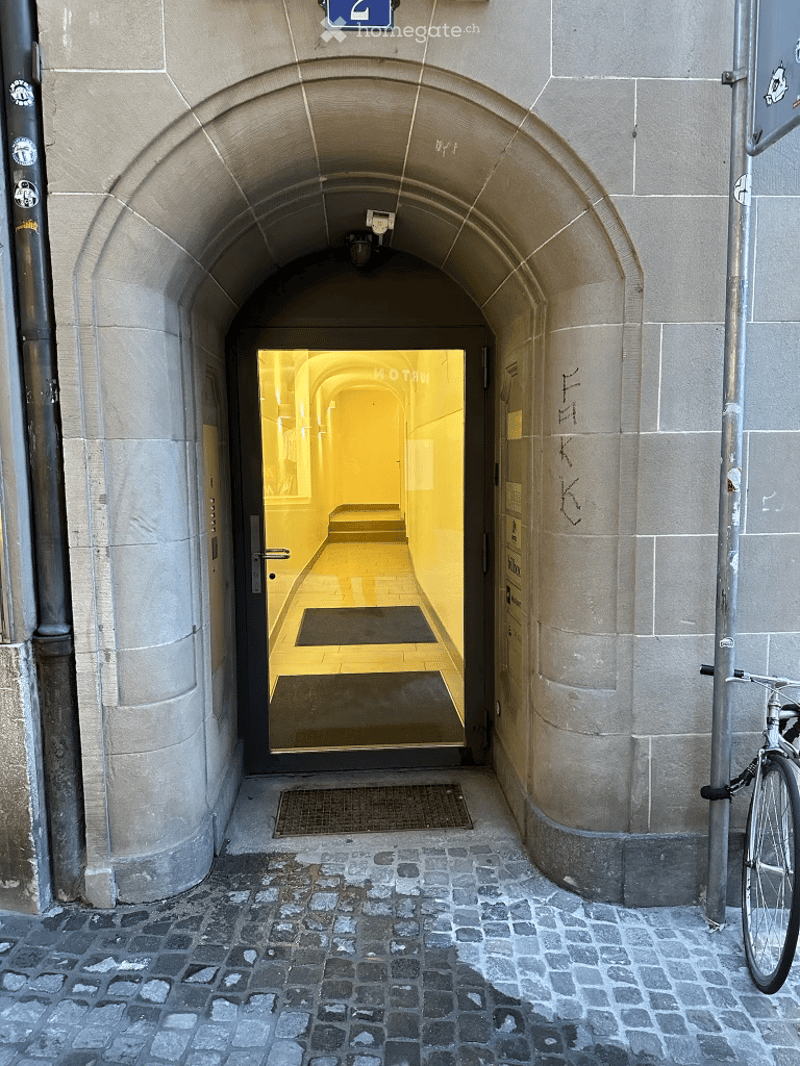 Entrance to a hallway with a stone archway, a glass door revealing a brightly lit interior corridor with a yellow-lit ceiling and tiled floor.