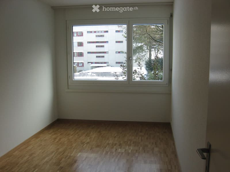 Empty room with wooden flooring and large windows showing a view of a building and trees outside.