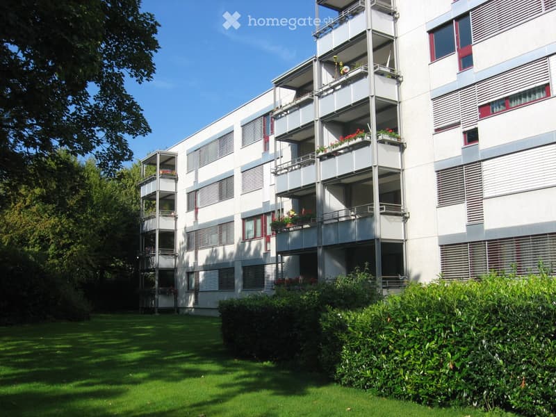 Modern multi-story apartment building with balconies, red-framed windows, and surrounding greenery.