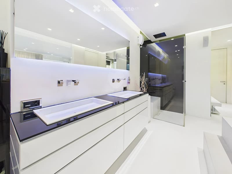 Modern bathroom with white cabinets, a large bathtub, a sink, and a large mirror. The space features sleek, contemporary design with integrated lighting and a glass-enclosed shower area visible in the background.