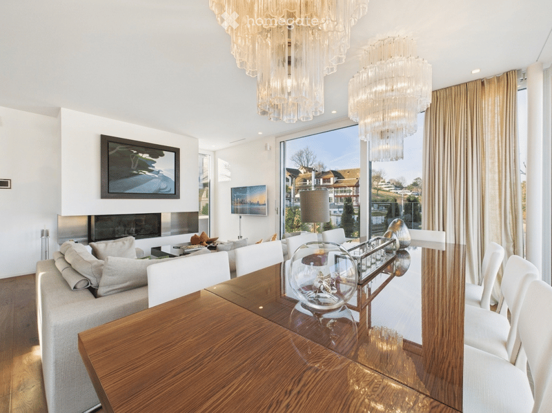 Modern living and dining area with large windows offering a scenic view, a grey sofa, a wooden dining table with white chairs, a fireplace, and decorative lighting.