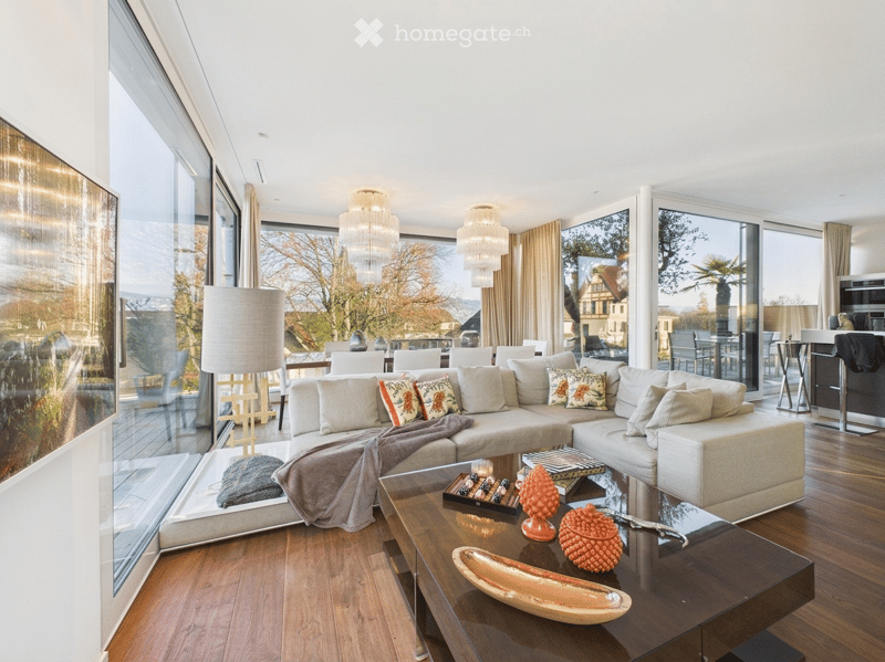 Modern living room with large windows offering a scenic view, featuring a sectional sofa, coffee table with decorative items, floor lamp, and additional seating.