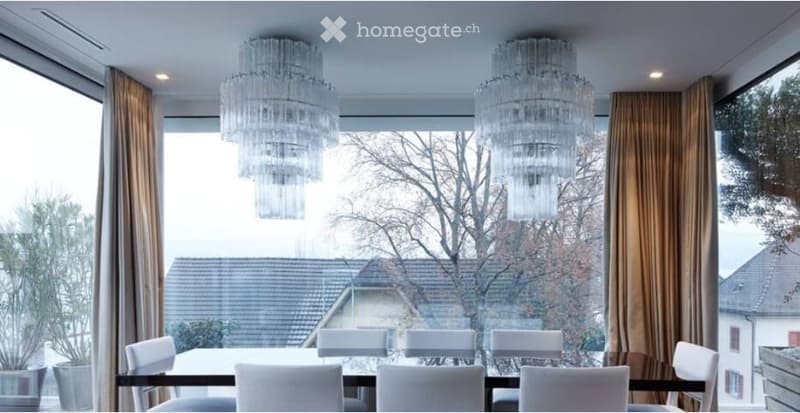 Elegant dining room with large windows offering a view of the surrounding area, featuring a dark wooden dining table surrounded by white chairs and two large crystal chandeliers.