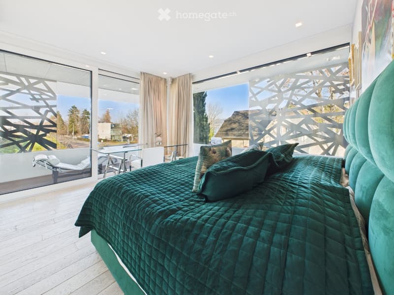 Modern bedroom with large windows offering a scenic view, a green duvet-covered bed, decorative pillows, a glass table, and sun loungers visible through the windows.