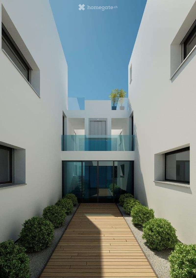 Modern outdoor courtyard with a wooden deck pathway, neatly trimmed shrubs, and a glass-enclosed balcony level above.