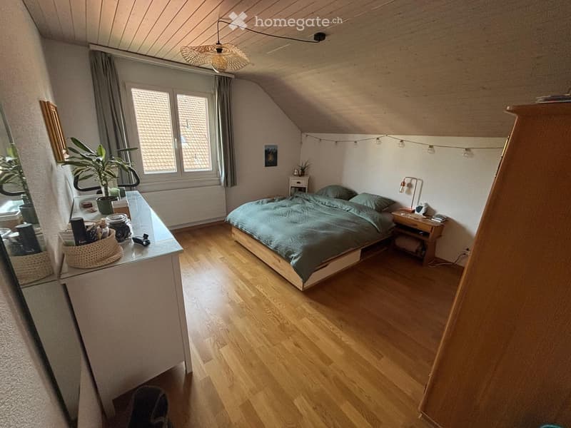Cozy attic bedroom with wooden floors, slanted ceilings, and large windows. Features a bed with green bedding, a nightstand with a lamp, a small table with a plant, and a mirror on the wall.