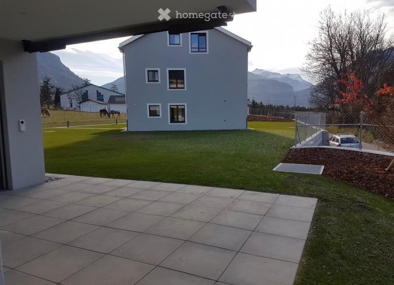 Outdoor patio area with tiled flooring, grassy lawn, and a modern house in the background. The scene includes a fenced area with a parked car and a view of mountains in the distance.