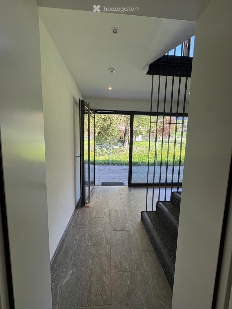 Modern hallway with marble flooring, white walls, and a staircase leading upwards. Large glass doors open to a garden area with greenery visible outside.
