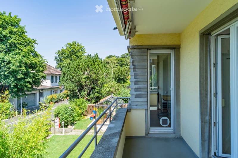 Balcony with metal railing, overlooking a green garden and neighboring house, with a clear view of the surrounding trees and a pathway.