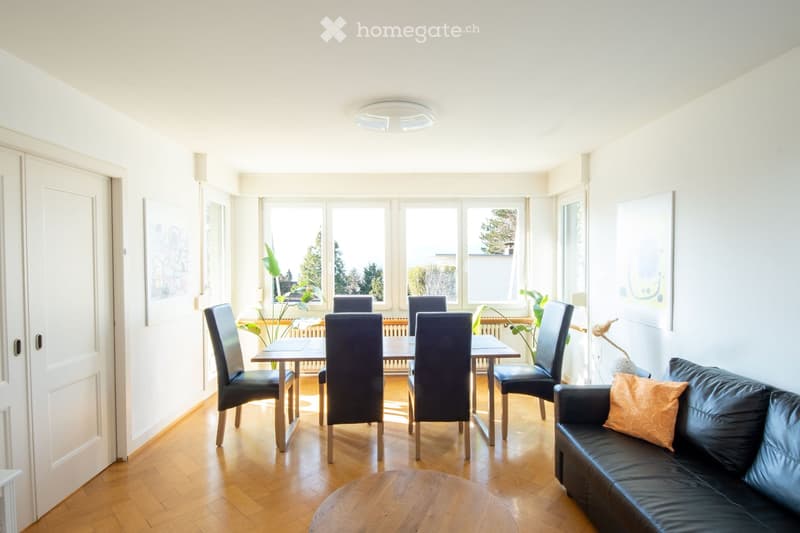 Bright living and dining area with wooden flooring, large windows, a black leather sofa, a wooden coffee table, a dining table with chairs, and a floor lamp.